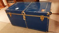 Front view of the blue metal clad wooden travel trunk with brass-tone latch and lock plate, showing light scratches and wear.
