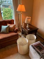 Photo showing one standing adjustable metal lamp with attached pleated fabric shade, another ceramic side table lamp on a wooden table with an attached stained fabric shade, along with three additional fabric lamp shades on the floor.