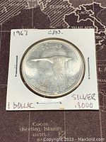Front side of the 1967 Canadian silver 1 dollar coin showing a flying Canada goose and coin details including '1 DOLLAR' and '1967'. The coin is inside a white cardboard holder with handwritten notes.
