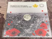 The commemorative coin displayed in a holder with historic military photo background and red poppies and maple leaves.