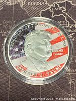Obverse side of silver coloured medallion showing profile of Donald Trump with US flag background and inscriptions.
