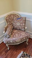 Front angle of armchair showing floral damask upholstery, carved wooden frame painted white, square back cushion and bolster pillow.