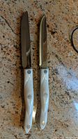 Two Cutco steak knives with white handles and serrated blades lying on granite countertop.