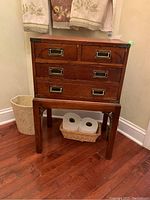 Front angled view of the wood storage chest with drawers and matching stand against wall with towels and basket beneath.