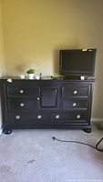 Full view of black dresser showing seven drawers and one cabinet door with items on top including TV, plant, decorative items