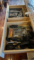 Drawer containing assorted flatware pieces, various spoons, forks, knives, and kitchen utensils including black plastic spatulas and ladles.