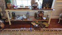 Full front view of sofa table with pottery, plates, glassware, and decorative items on two shelves, placed on wooden floor with patterned rug in front.