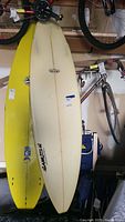 Full view of the beige surfboard leaning against a wall, showing brand logos and general shape
