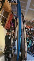 Full side view of the blue and white Riviera surfboard stored vertically among garage items.