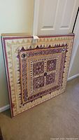 Framed square tapestry fabric art with geometric and decorative patterns leaning against a wall.