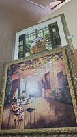 Two stacked framed wall art pieces, showing floral window painting above and outdoor patio scene below