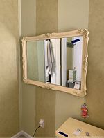 Wall mirror with cream colored ornate frame hung on beige wall, showing floral and scroll details. Visible chipping on frame edges.