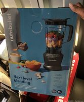 Box front showing NutriBullet blender with pitcher filled with fruits, highlighting 1200 watt motor base, 64 oz blender pitcher, BPA free, dishwasher safe, pulse function, and recipe book included.