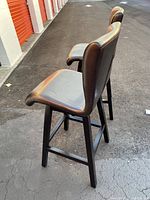 Two dark brown ultrahyde upholstered barstools viewed from side and slightly diagonal angles, showing wooden legs and footrests, curved backrests, and cushioned seats.