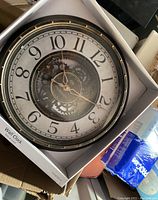 Wall clock in box, showing clock face with black numbers and visible bronze gears in center. Clock measures 39cm diameter.
