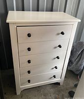 Front view of white wooden chest with five drawers and black round knobs, showing the drawer fronts and top surface.