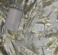 White Amazon Echo 3rd Gen smart speaker and white power adapter with cable on patterned fabric background, showing front and side of speaker and adapter cable arrangement.