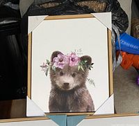 Framed wall art featuring a realistic baby bear wearing a purple flower crown, resting on a beige surface with protective corner covers attached.