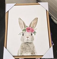 Photo of one framed baby bunny rabbit painting with a pink floral crown, protected by white corner covers and a wooden frame, displayed upright.