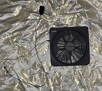 Front view of battery operated electric fan showing circular fan grill and attached power cord with battery pack.