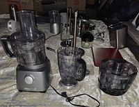 Photo showing silver base unit with control dial, 8-cup food processor bowl, 40 oz blender jar, and continuous juice extractor bowl with lids and blade parts spread out on surface.