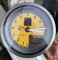 Photo shows a round wall clock with a stainless steel rim and a yellow and gray tachometer-styled dial including Ferrari prancing horse logo, marked rpm x1000, with digital display windows and clock hands.