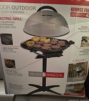 Box front showing the George Foreman Indoor/Outdoor Electric Grill with 15 servings capacity, variable temperature control, nonstick coating, removable stand, and unique sloped surface for fat drainage.