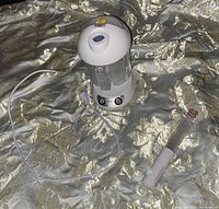 Hair and facial steamer showing transparent water tank, control knobs, power cord, and detachable nozzle/cover piece placed beside it on fabric surface.