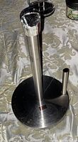 Vertical and angled views of stainless steel paper towel holder with weighted base on patterned tablecloth.