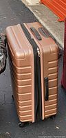 Side view of the rose gold large hard shell luggage bag standing on floor showing the black handle and zipper.