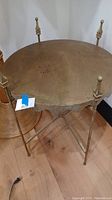 Overall front view of a round brass side table with fold-up design, four slender corner posts with finials, scratched top, and scalloped edge around tabletop.