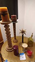 View of two tall carved wooden pillar candle holders with candles, one brass palm tree candle holder with candle, and several loose candles arranged on a wooden table.