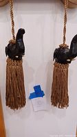 Pair of curtain tiebacks with dark carved elephant heads and brown tassels hanging on wall