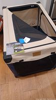 Large beige and black precision soft side pet crate showing zip open mesh window and overall structure on wooden floor.