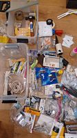 Overview of assorted tools and hardware items spread on floor including saw, hammer, electrical parts, tapes, cords, and packaging.