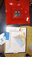 Two trays shown together. One is red Murano glass with four colorful glass cubes in the center. The other is a white ceramic rectangular tray with a painted gargoyle design and signature tape tags attached.