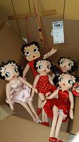 Four Betty Boop dolls inside a cardboard box, including a marionette with wooden control bars and three plush dolls in red and pink dresses.