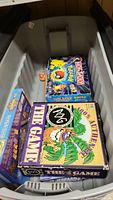 Bin containing assorted board games including Pokémon Monopoly and The Game, showing top view inside bin.
