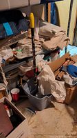 Full view of shelf and floor showing painting supplies including gray plastic bucket with paint roller extension pole, drop cloths, boxes, and other smaller items