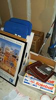 Photo showing blue and white folding sports chairs leaning against a wall, a box of baseball cards, and framed posters stacked
