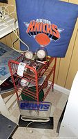 Photo showing the red metal sports rack containing baseballs in protective cases, New York Knicks pillow, and New York Giants lighted plaque on the base.