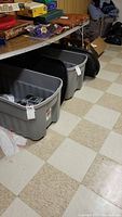 Photo showing two large gray Rubbermaid Roughneck storage bins with black lids placed under a table in a basement area with tiled floor and various items on the table above.