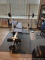 Assembled bench press setup with padded bench, barbell with weights, and support rack in room with window and mat flooring.