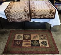 Two small rugs with brown-beige designs hanging side by side on a table with a third rug in earth tones featuring leaf patterns laid on the floor.