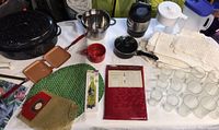 Photo showing a variety of kitchen and dining items including black turkey roaster, metal strainer, copper-colored sandwich maker, red mugs, water filter pitchers, small black saucepan, and stack of linens.
