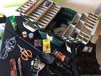 Photo showing fishing tackle box opened with compartments of fishing supplies, fishing rod, tactical pack, scissors, knife, insect repellent, and various smaller items arranged on table.