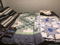 Photo shows multiple folded quilts and throws laid out on beds or furniture including striped and floral patterned pieces with two packs of Eclipse blackout curtains.