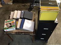 Full view showing 3 drawer black locking file cabinet with key and portable hanging folder frame with folders on top. Stack of ring binders and sheet protector box to the side.