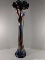 Tall iridescent glass vase with ruffled top and ribbed texture, standing on a white surface against a neutral background.