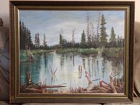 Framed landscape painting featuring a lake with trees and reflections, wood frame with visible wear.